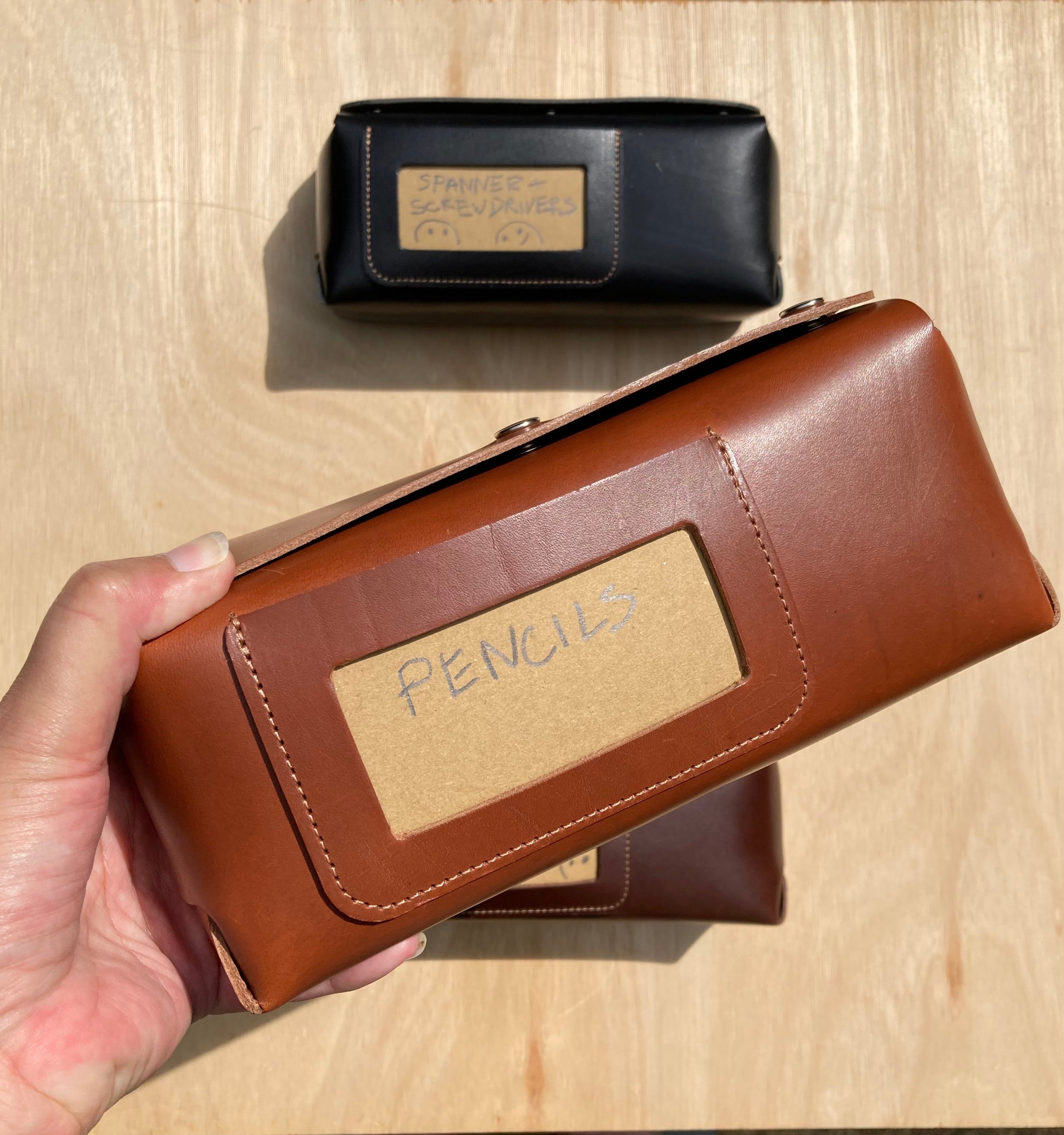 Brown pencil case with 'Pencils' label held by a hand on a wooden surface
